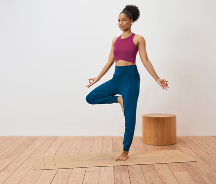 Frau in pinkem Top und blauer Hose steht in der Baumposition auf einer Yogamatte.