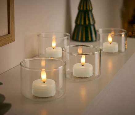Vier weiße Kerzen in durchsichtigen Glasbehältern mit einem kleinen grünen Weihnachtsbaum im Hintergrund.
