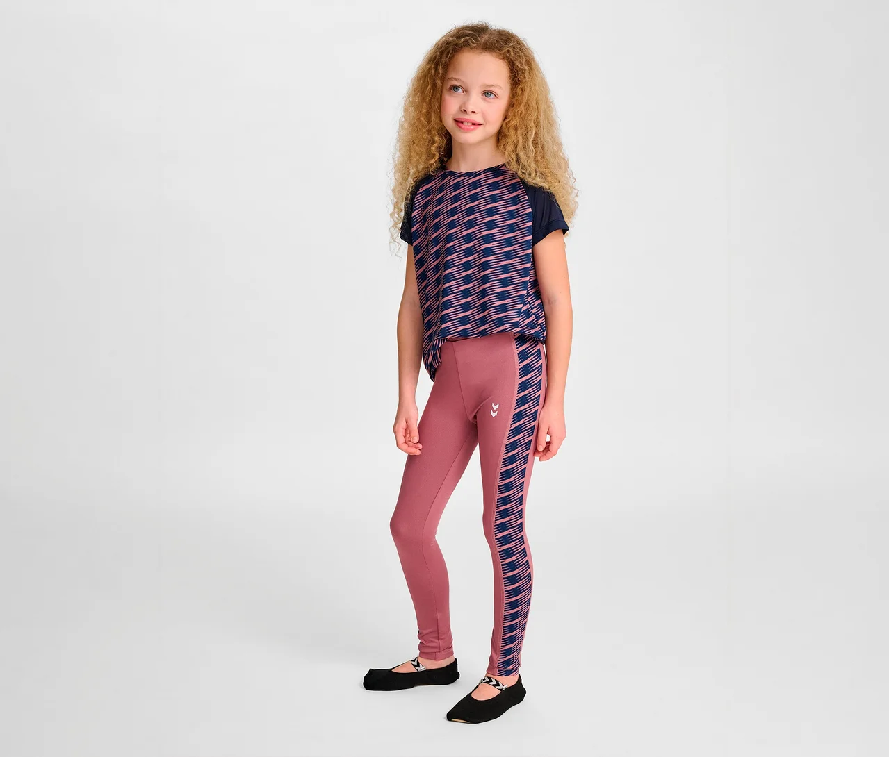 Mädchen steht und trägt ein geometrisch gemustertes T-Shirt und eine rosafarbene HUMMEL HMLJR Mix Mid Waist Tight Leggings.