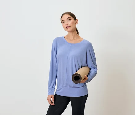Frau hält eine zusammengerollte Yogamatte, trägt ein blaues Langarmshirt und schwarze Leggings.