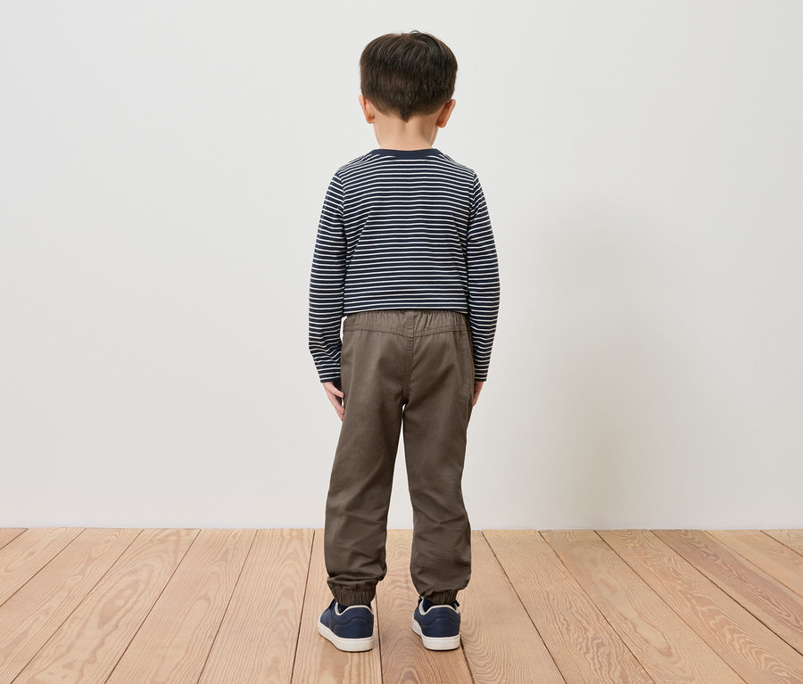 Ein Junge steht mit dem Rücken zur Kamera und trägt ein gestreiftes Shirt, braune Hosen und blaue Schuhe.