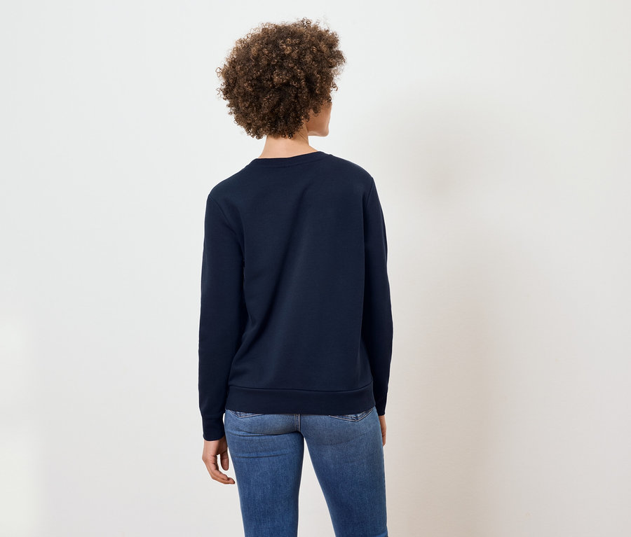 Frau mit lockigem Haar steht mit dem Rücken zur Kamera und trägt ein marineblaues Sweatshirt und Jeans.