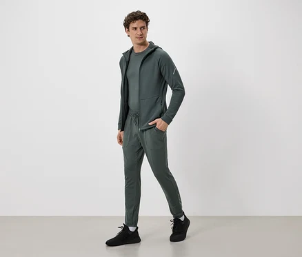 Mann in olivgrünem Trainingsanzug mit Kapuzenjacke und Hose mit Kordelzug, dazu schwarze Turnschuhe.