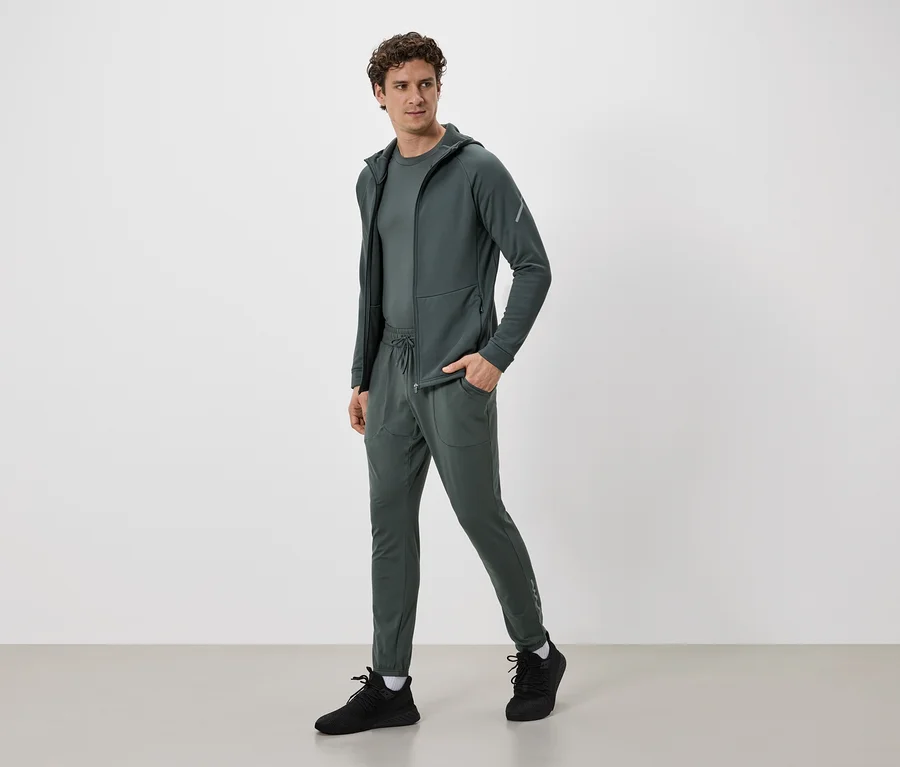 Mann in olivgrünem Trainingsanzug mit Kapuzenjacke und Hose mit Kordelzug, dazu schwarze Turnschuhe.