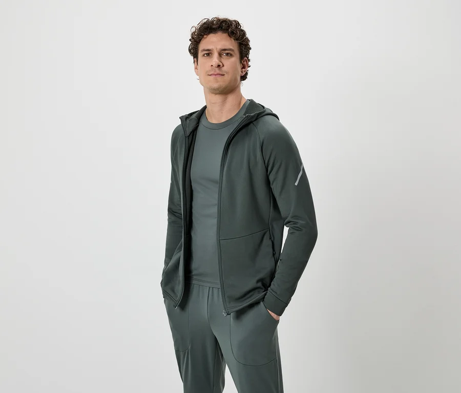 Mann mit lockigem Haar, trägt ein dunkelgrünes Trainingsanzug-Set, bestehend aus Kapuzenjacke, T-Shirt und Jogginghose, vor einem neutralen Hintergrund.
