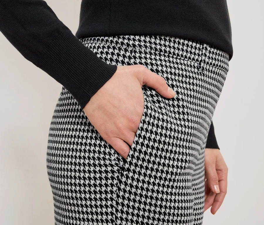 Frau mit Hand in der Tasche einer schwarz-weiß karierten Hose und einem schwarzen Pullover.