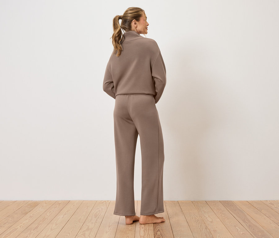 Eine Frau steht mit dem Rücken zur Kamera und trägt einen braunen Hosenanzug.
