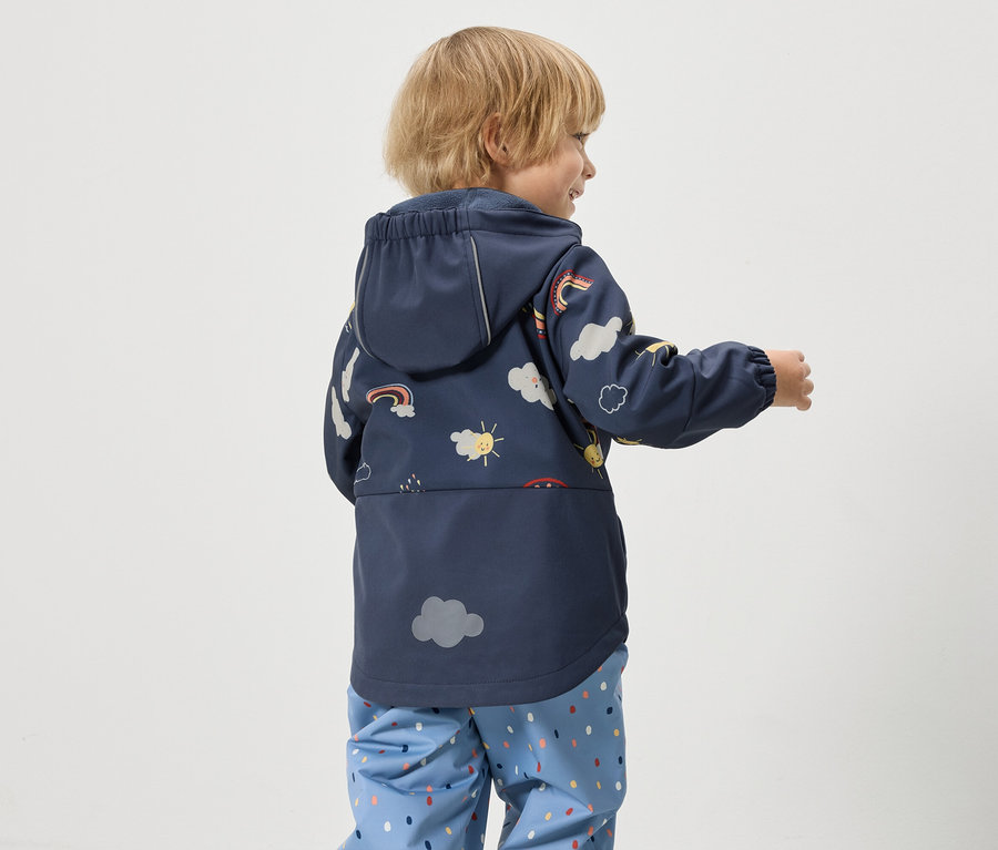 Ein Kind trägt eine dunkelblaue Regenjacke mit Kapuze, die mit Wolken, Sonnen und Regenbögen bedruckt ist, sowie blaue Regenhosen mit bunten Punkten.