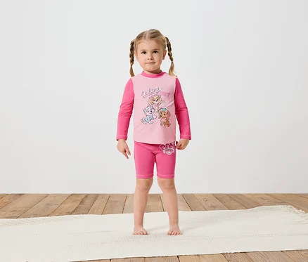 Ein kleines Mädchen mit Zöpfen steht barfuß auf einem cremefarbenen Teppich und trägt ein rosafarbenes Paw Patrol-Langarmshirt und Shorts.