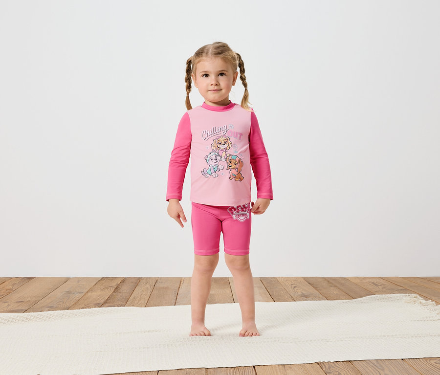 Ein kleines Mädchen mit Zöpfen steht barfuß auf einem cremefarbenen Teppich und trägt ein rosafarbenes Paw Patrol-Langarmshirt und Shorts.