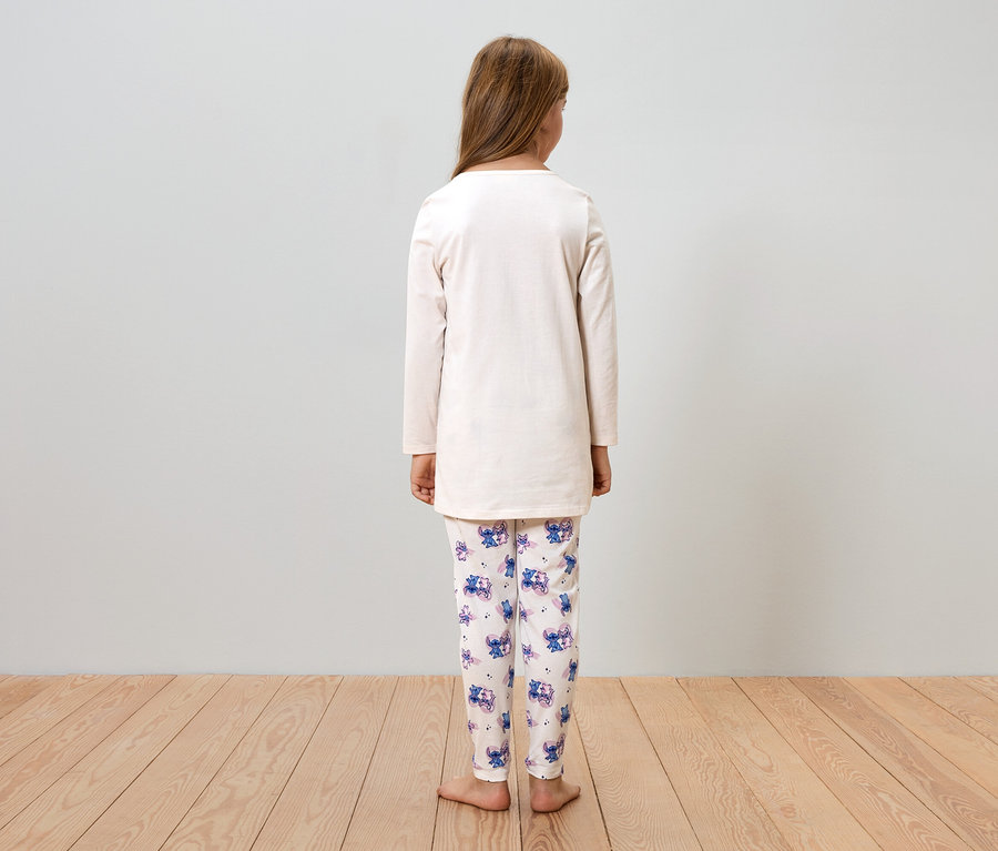 Mädchen von hinten, trägt einen beige Pyjama mit Stitch-Motiven, steht auf einem Holzboden vor einer grauen Wand.