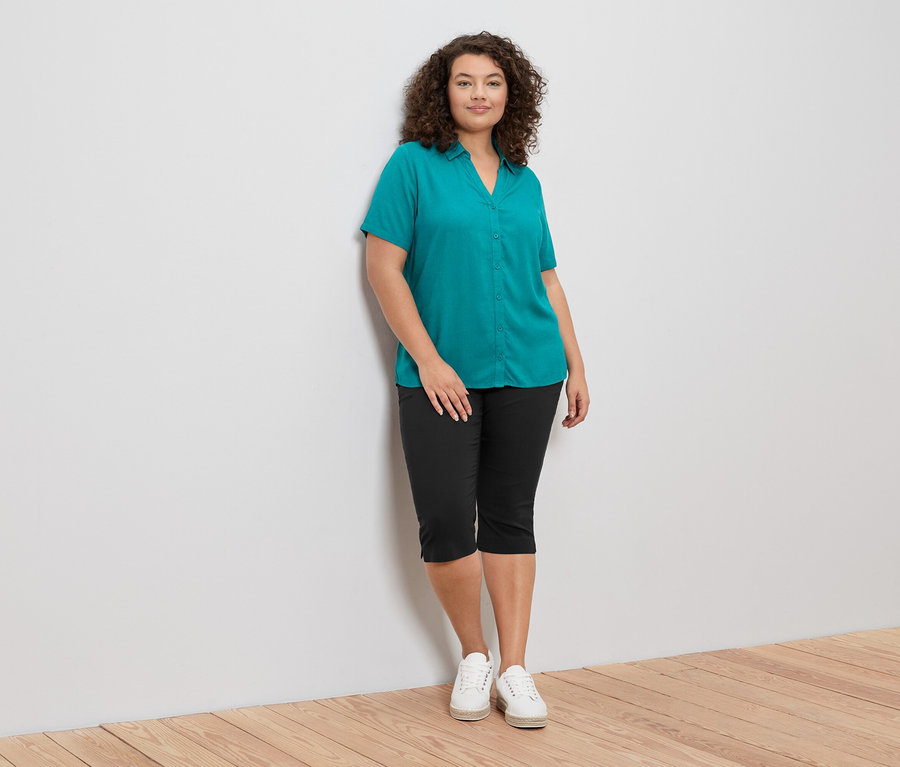 Frau in grüner Bluse und schwarzer 3/4-Bengalin-Hose. Dazu trägt sie weiße Schuhe.