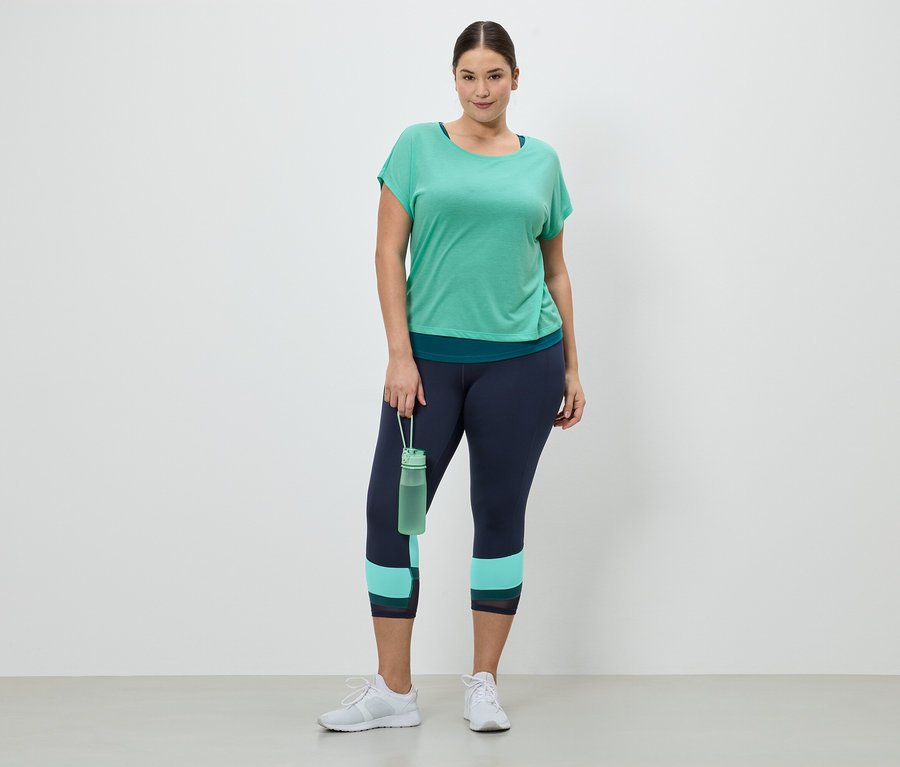 Frau in Sportbekleidung, bestehend aus einem grünen T-Shirt, einer marineblauen Leggings und weißen Turnschuhen, hält eine grüne Wasserflasche.
