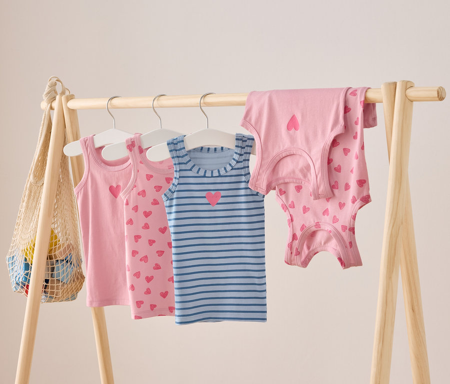 An einer Holzstange hängen rosa und blau gestreifte Kinder Tank Tops mit Herz Motiven.
