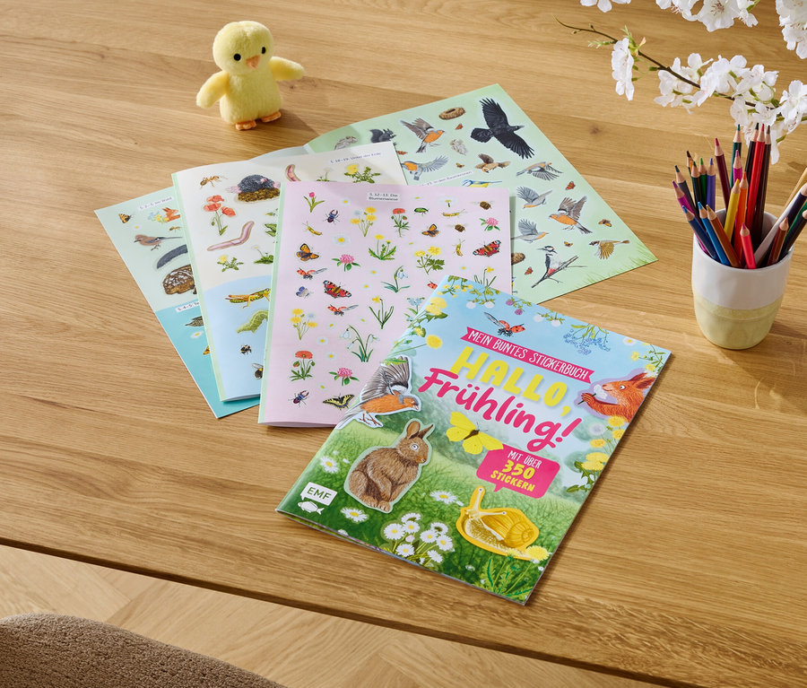 Auf einem Holztisch liegen ein gelbes Küken, Stickerbücher und Buntstifte in einem Becher.