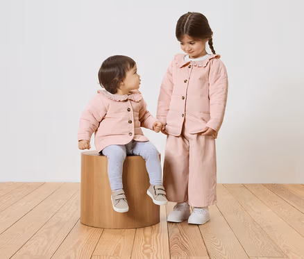 Zwei Mädchen, eines sitzt auf einem Holzhocker, beide tragen rosa Jacken mit Rüschenkragen.