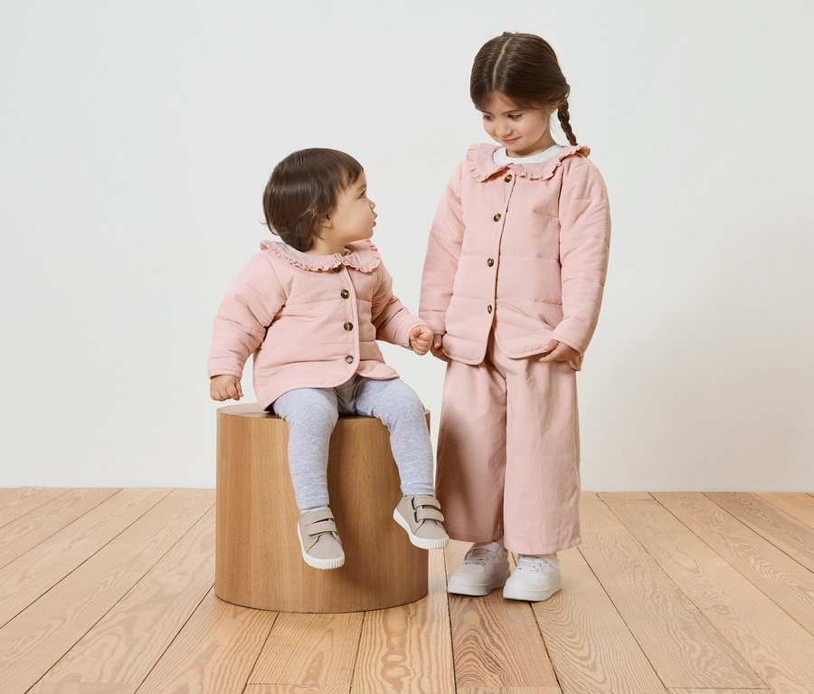 Zwei Mädchen, eines sitzt auf einem Holzhocker, beide tragen rosa Jacken mit Rüschenkragen.