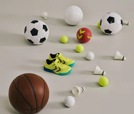 Gelbe hummel® Multiplay Flex VC JR Schuhe zwischen Bällen und Federbällen auf hellem Grund.