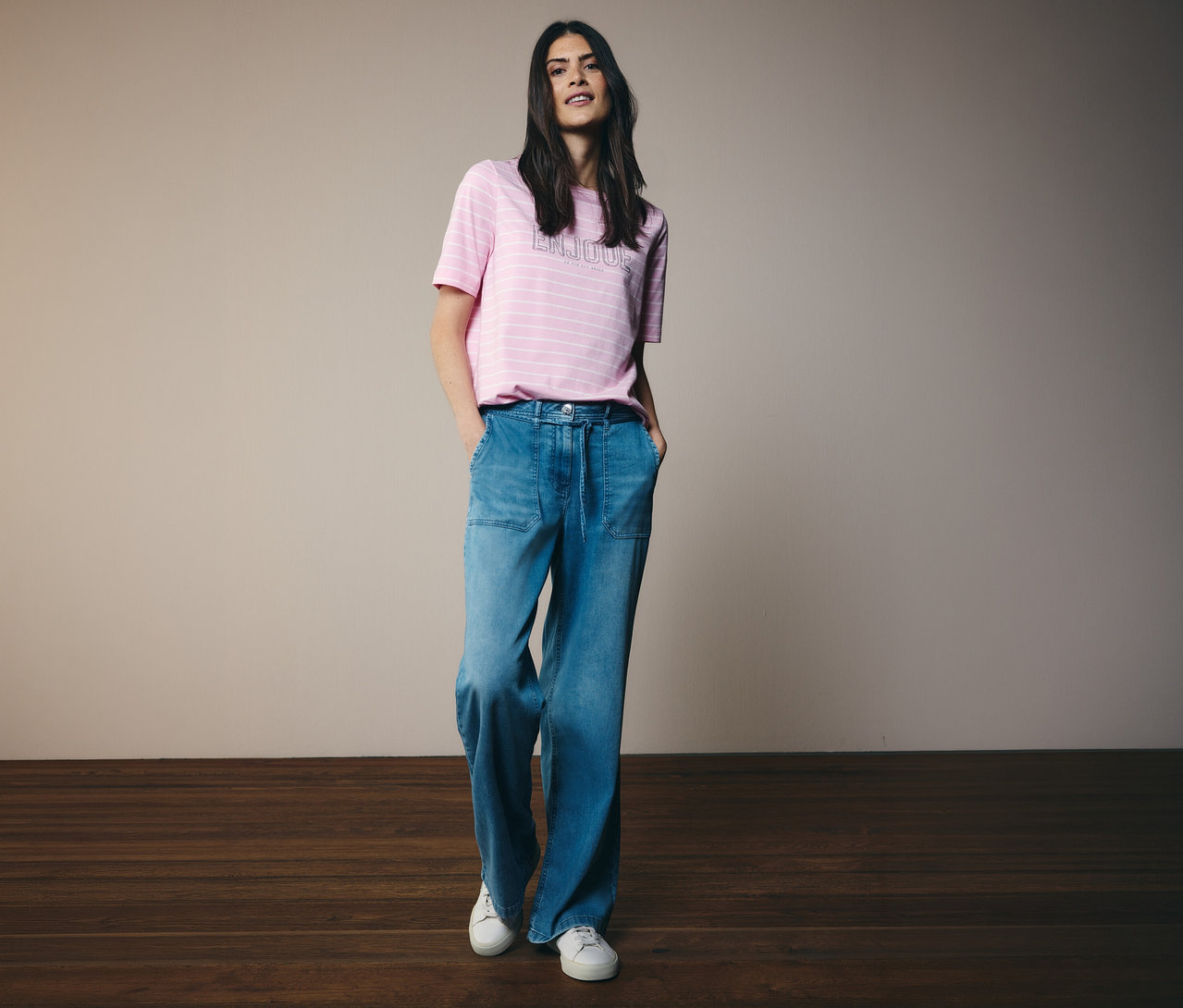 Frau mit CECIL Shirt mit rosa Streifen und Jeans mit weitem Bein und weißen Turnschuhen, die mit den Händen in den Hosentaschen posiert.