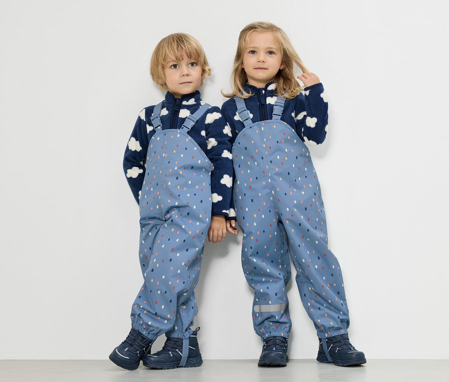 Zwei Kinder stehen nebeneinander, ein Junge und ein Mädchen, beide tragen blaue Fleecejacken mit Wolkenmuster und blaue Regenlatzhosen mit bunten Punkten.