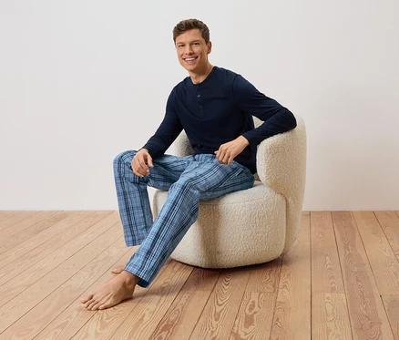 Ein Mann sitzt barfuß in einem Sessel, er trägt einen dunkelblauen Pullover und eine blau karierte Hose.