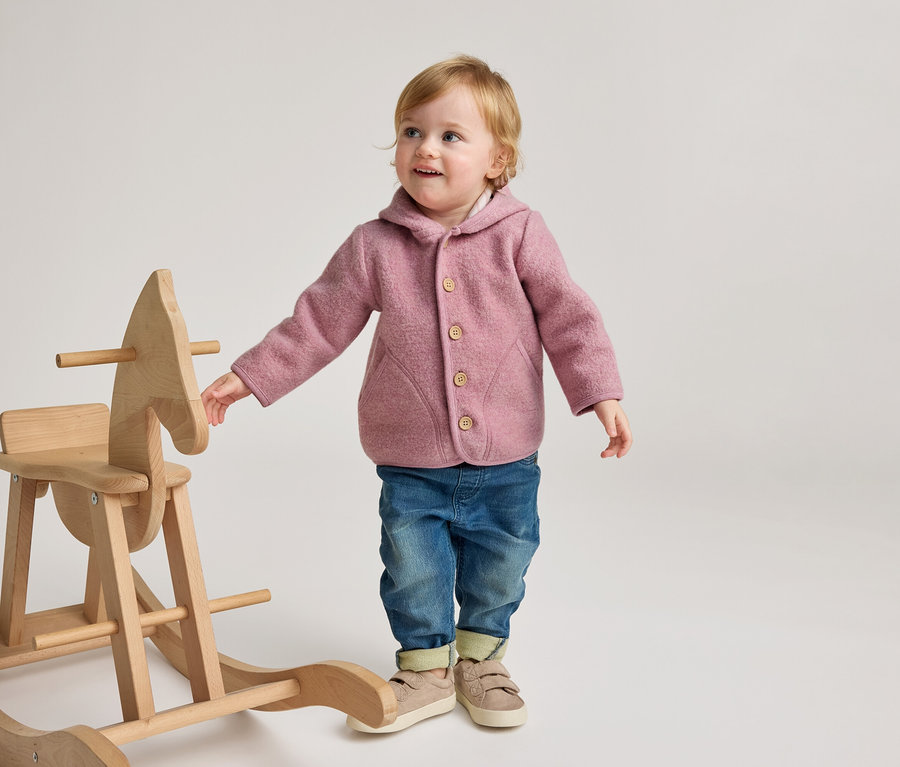 Ein lächelndes Kleinkind in einer rosa Kapuzenjacke, blauen Jeans und hellen Schuhen steht neben einem hölzernen Schaukelpferd und blickt mit einer Hand am Pferd nach oben.