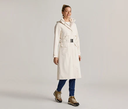 Frau in einem langen, cremefarbenen Mantel mit Gürtel, blauen Jeans und braunen Stiefeln.