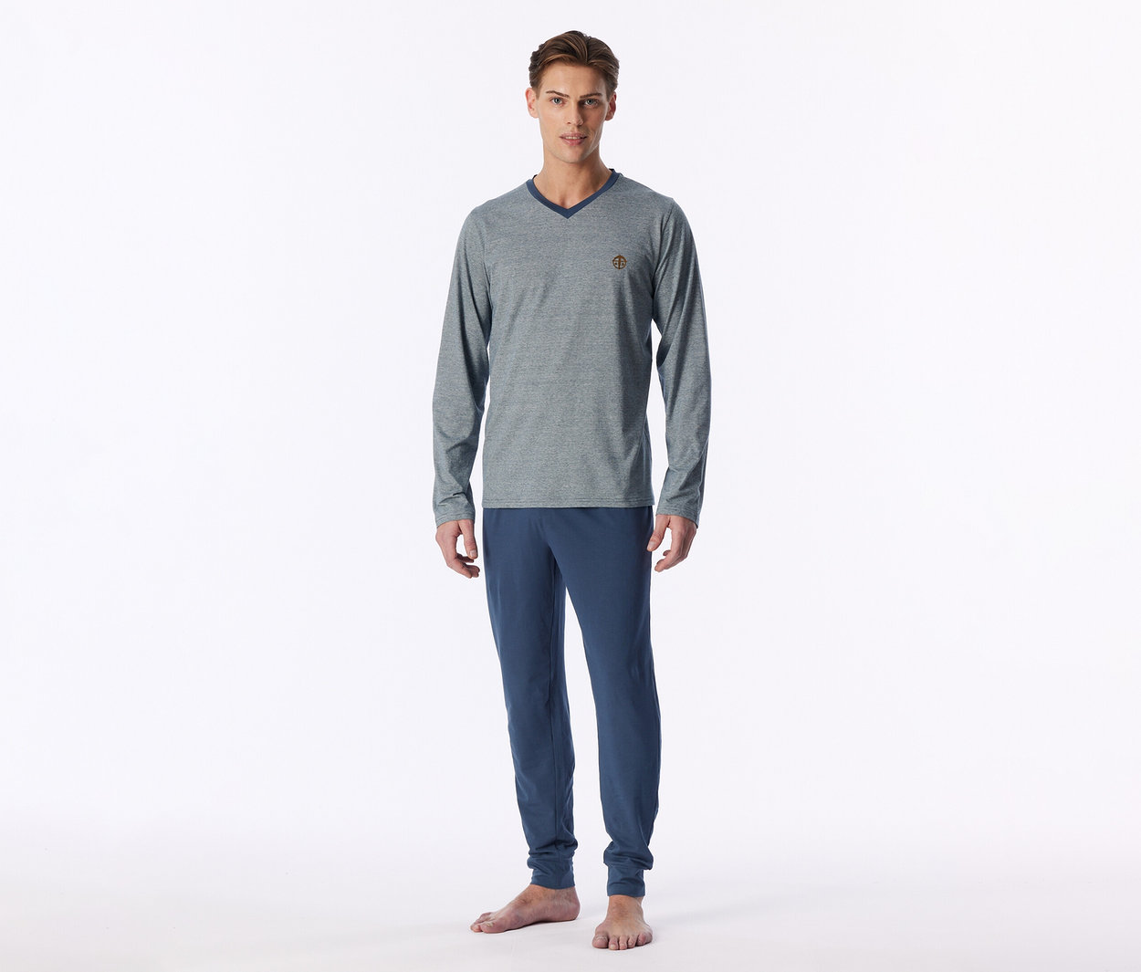 Mann steht barfuß und trägt einen grauen, langärmligen V-Ausschnitt-Pullover mit dunkelblauem Kragen und eine blaue Jogginghose.