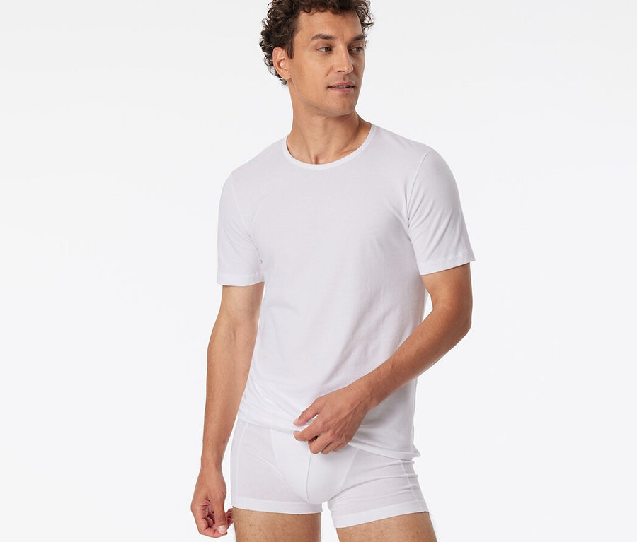Mann mit lockigem Haar trägt ein weißes SCHIESSER T-Shirt und weiße Boxershorts, blickt nach rechts und zieht den Saum seines T-Shirts hoch.