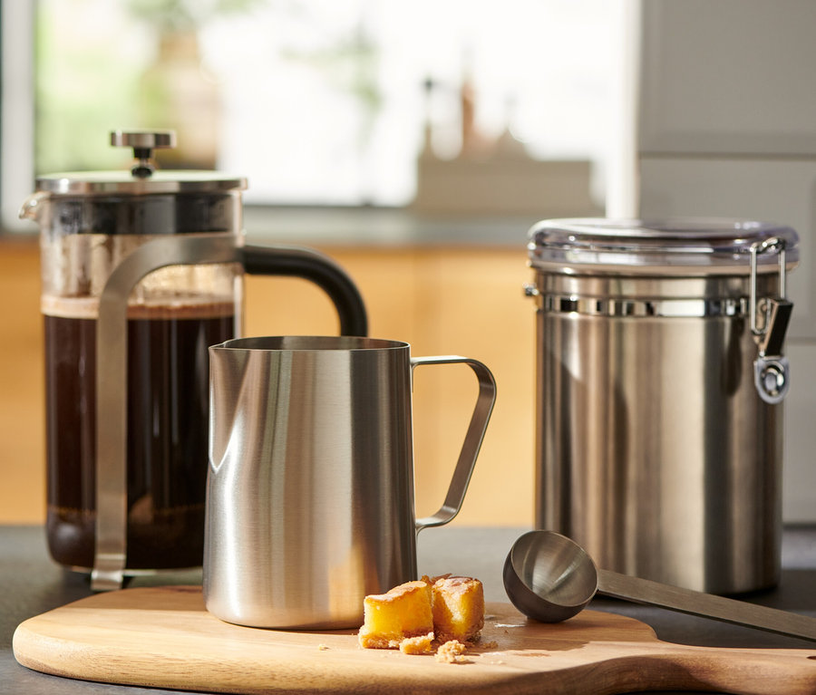Ein Glas Kaffee steht neben einer French Press und einem kleinen Milchkännchen aus Edelstahl auf einem Holzbrett.