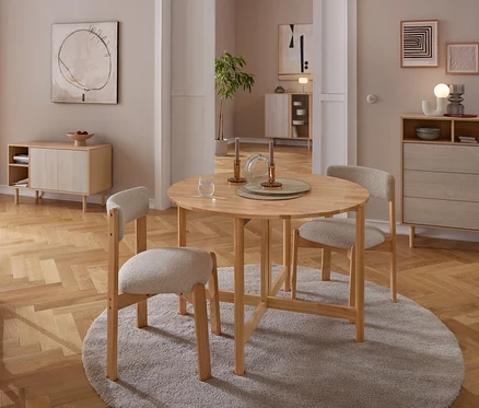 Ein Essbereich mit einem runden Holztisch und drei hell gepolsterten Stühlen auf einem grauen Teppich. Der Tisch ist mit Kerzenständern und einem Glas gedeckt. Im Hintergrund stehen ein Holz-Sideboard und eine Kommode an neutralen Wänden mit abstrakter Kunst, alles auf Parkettboden.