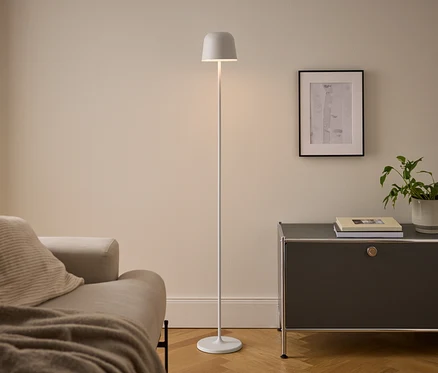 Ein modernes Wohnzimmer mit beigem Sofa, einer weißen Stehlampe und einem dunkelgrauen Schrank. Die beleuchtete Lampe steht zwischen Sofa und Schrank, auf dem Bücher und eine Topfpflanze unter einem gerahmten Bild liegen.