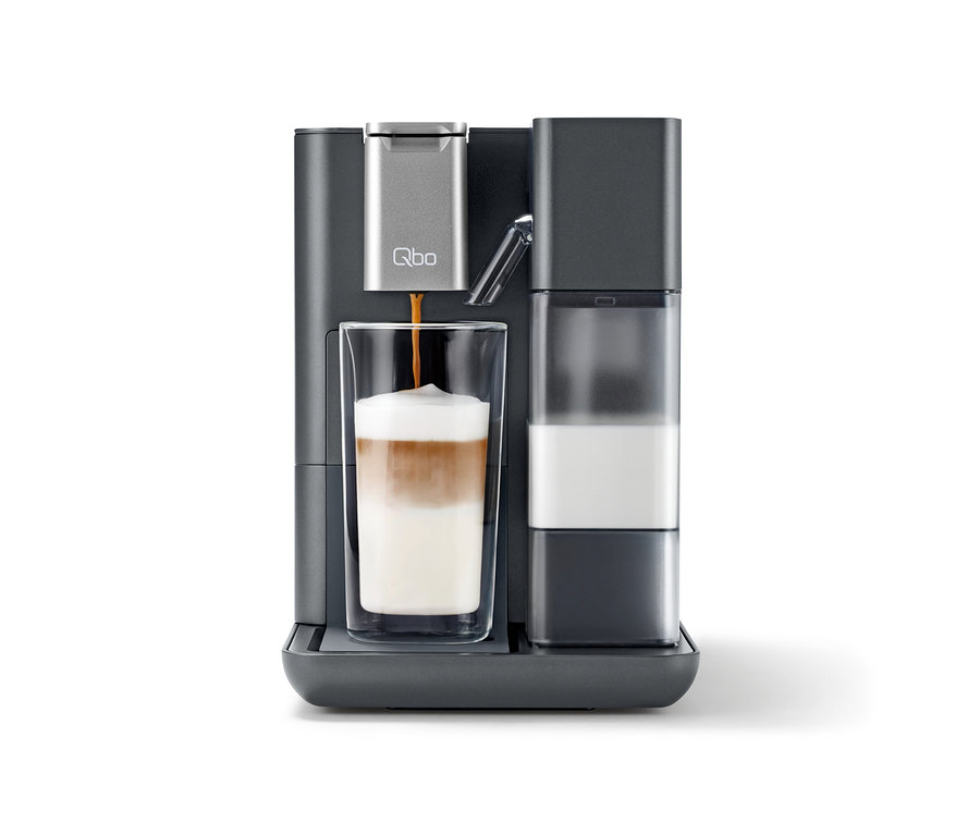 Dunkelgraue Qbo MILK MASTER Gentle Grey Kaffeemaschine, die Kaffee in ein hohes Glas mit geschichtetem Latte Macchiato füllt, daneben ein Milchbehälter.