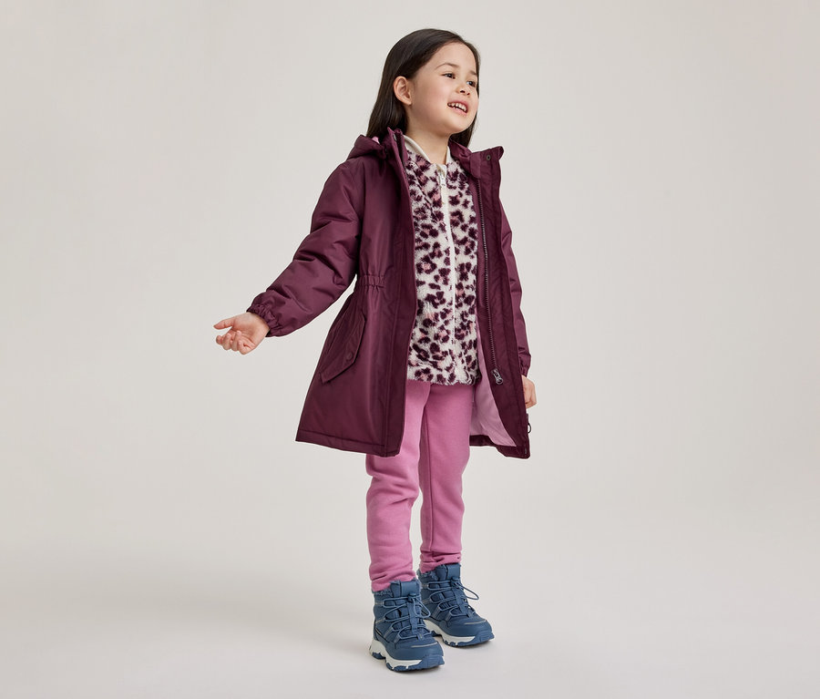 Ein junges Mädchen steht lächelnd und blickt nach rechts. Sie trägt einen pflaumenfarbenen Kapuzenparka über einer weiß-lila Fleecejacke mit Leopardenmuster, rosa Hosen und blaue Stiefel.