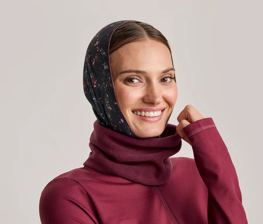 Lächelnde Frau mit dunklem, geblümtem Kopftuch und bordeauxrotem Rollkragenpullover, die den Kragen hält.