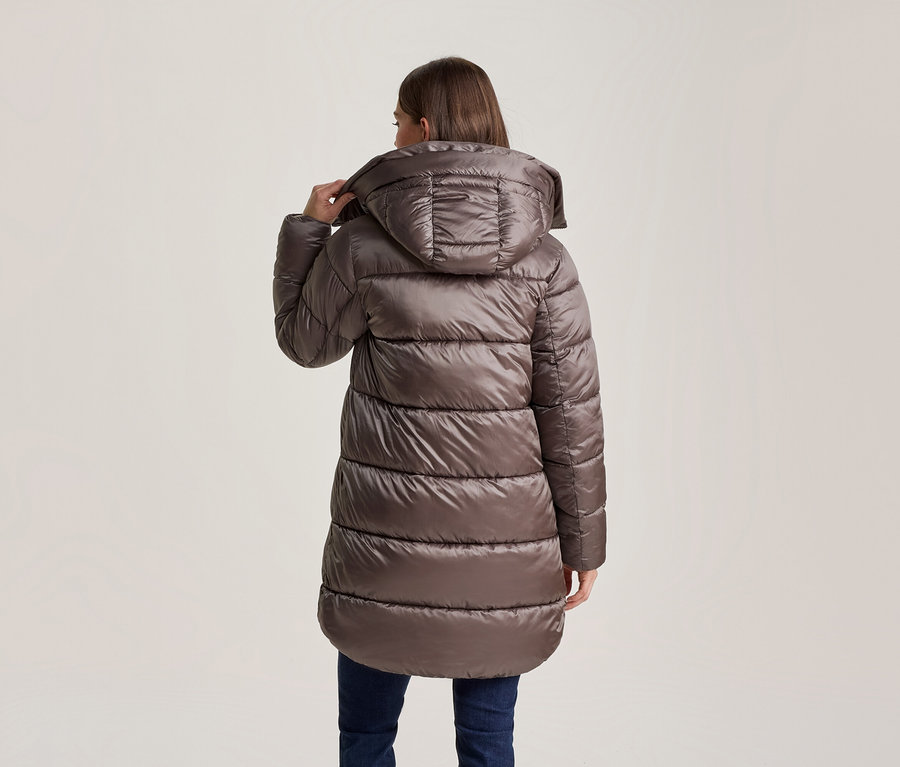 Frau von hinten in einer langen, braunen Steppjacke mit Kapuze, die sie mit der Hand anpasst, dazu dunkle Jeans.