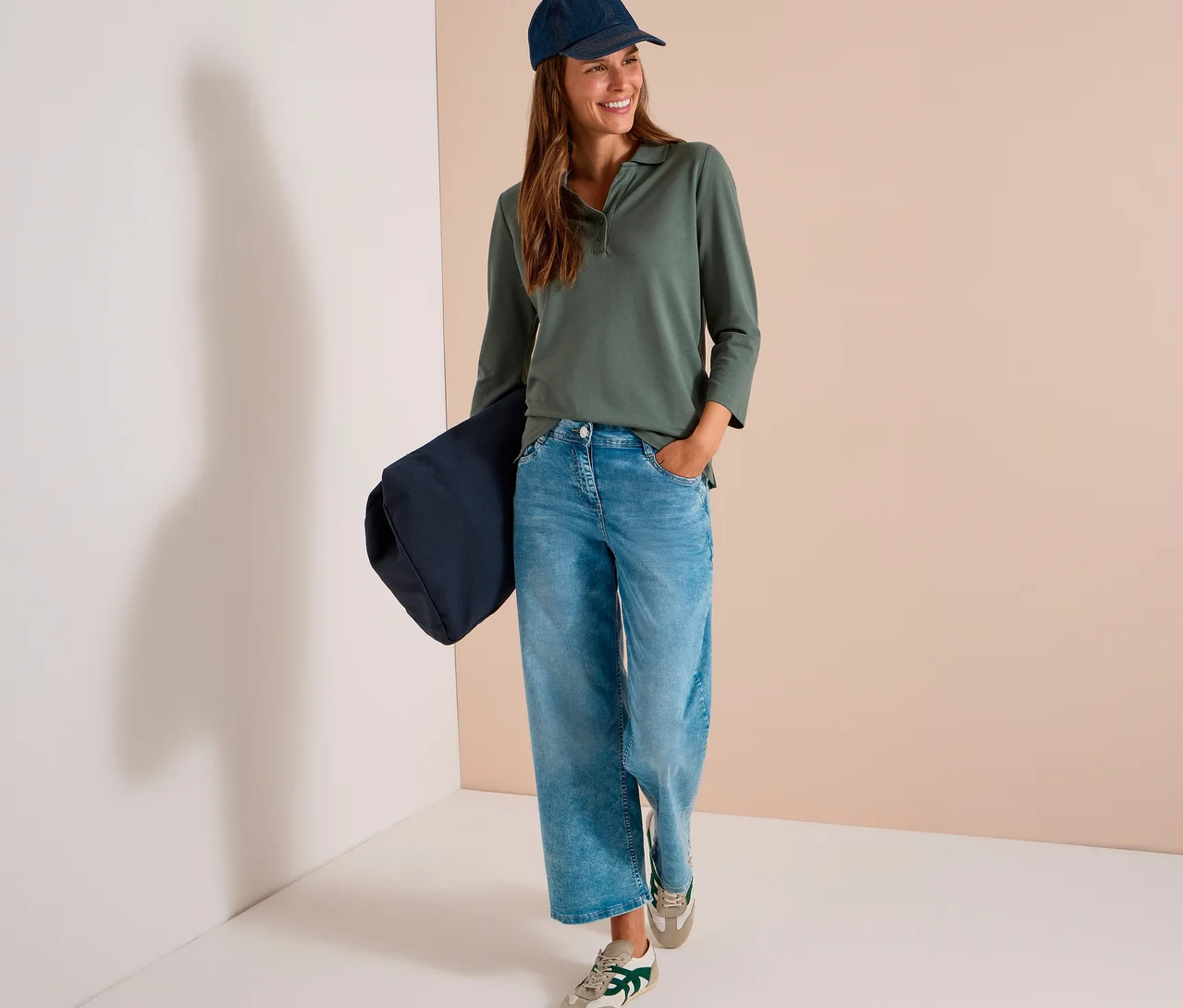 Lächelnde Frau trägt ein grünes CECIL 3/4-Poloshirt, hellblaue Jeans, eine dunkelblaue Kappe und Turnschuhe, eine dunkelblaue Tasche haltend.