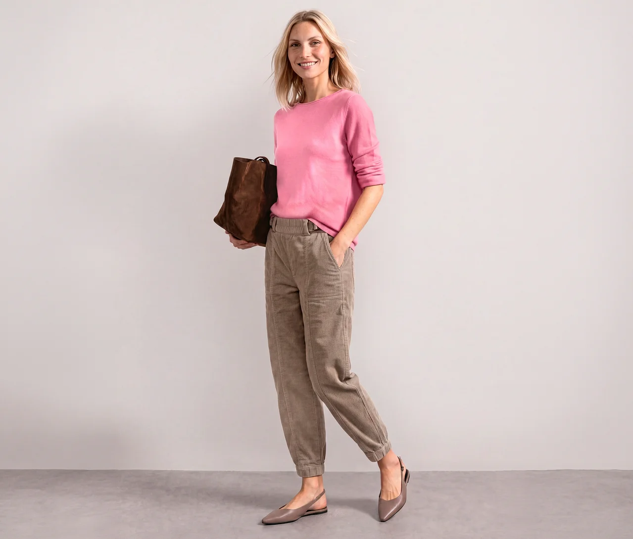 Lächelnde Frau in einem rosafarbenen Street One Strickpullover, hellbraunen Hosen und taupefarbenen Flats, eine braune Tasche haltend.