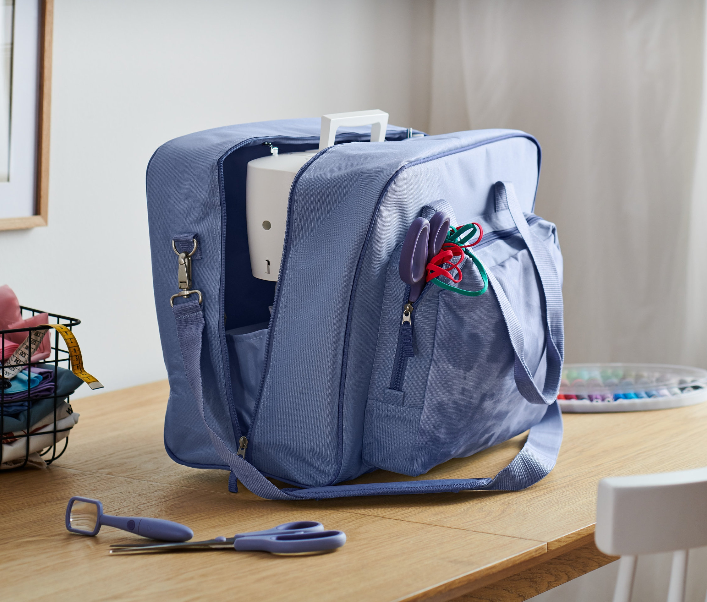 Eine blaue Nähmaschinentasche steht offen auf einem Holztisch, im Hintergrund ein Stoffkorb und ein Fenster.