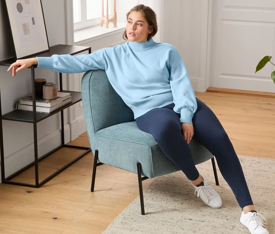 Eine Frau sitzt in einem Sessel und trägt Sneaker aus chromfrei gegerbtem Leder, Leggings und einen Pullover mit Ballonärmeln.