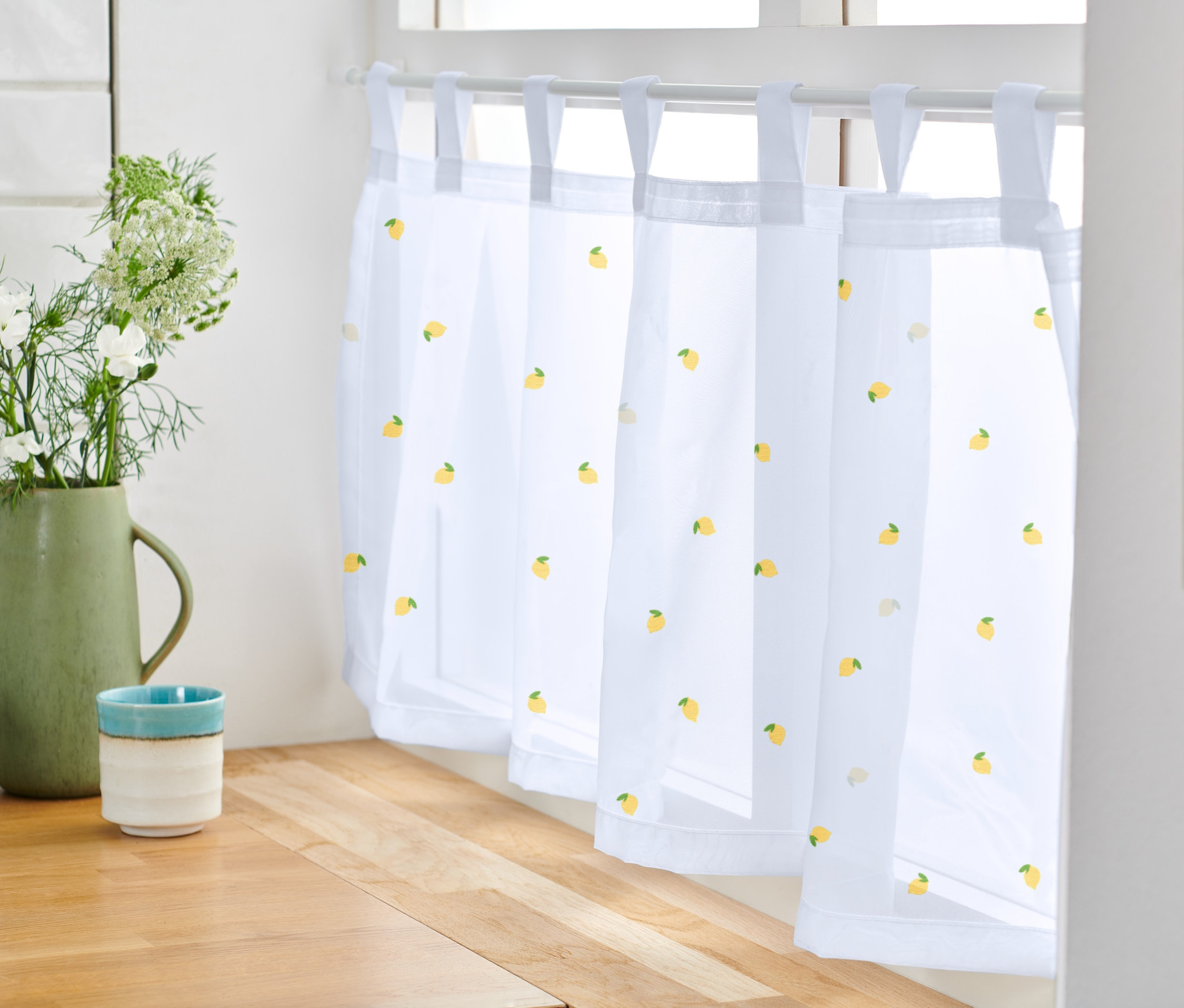 Eine bestickte Bistro-Gardine mit Zitronenmuster hängt am Fenster. Daneben stehen eine Vase mit Blumen und eine Tasse.