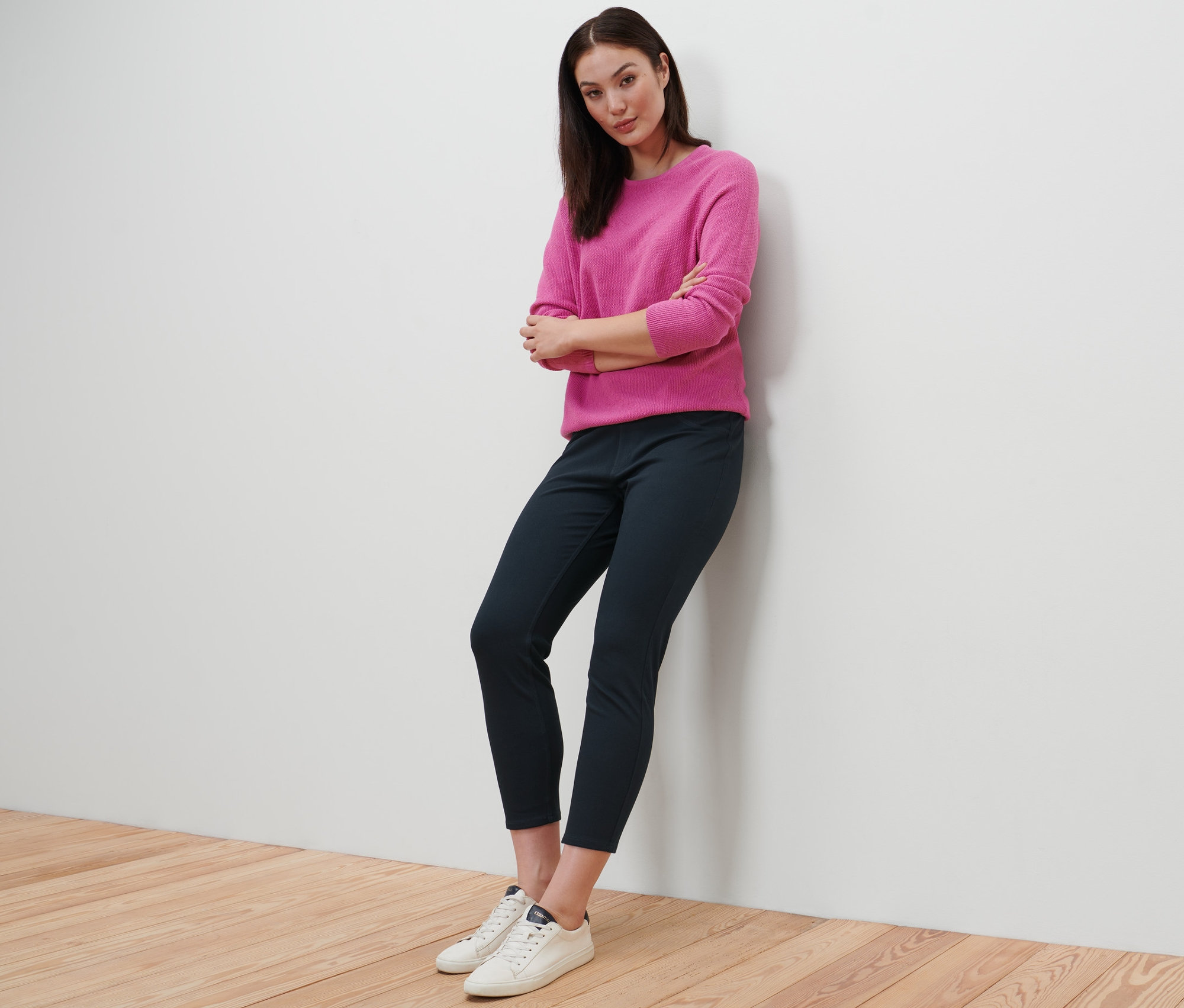 Eine Frau lehnt an einer Wand und trägt eine 7/8-Jeggings, einen pinken Pullover und weiße Turnschuhe.