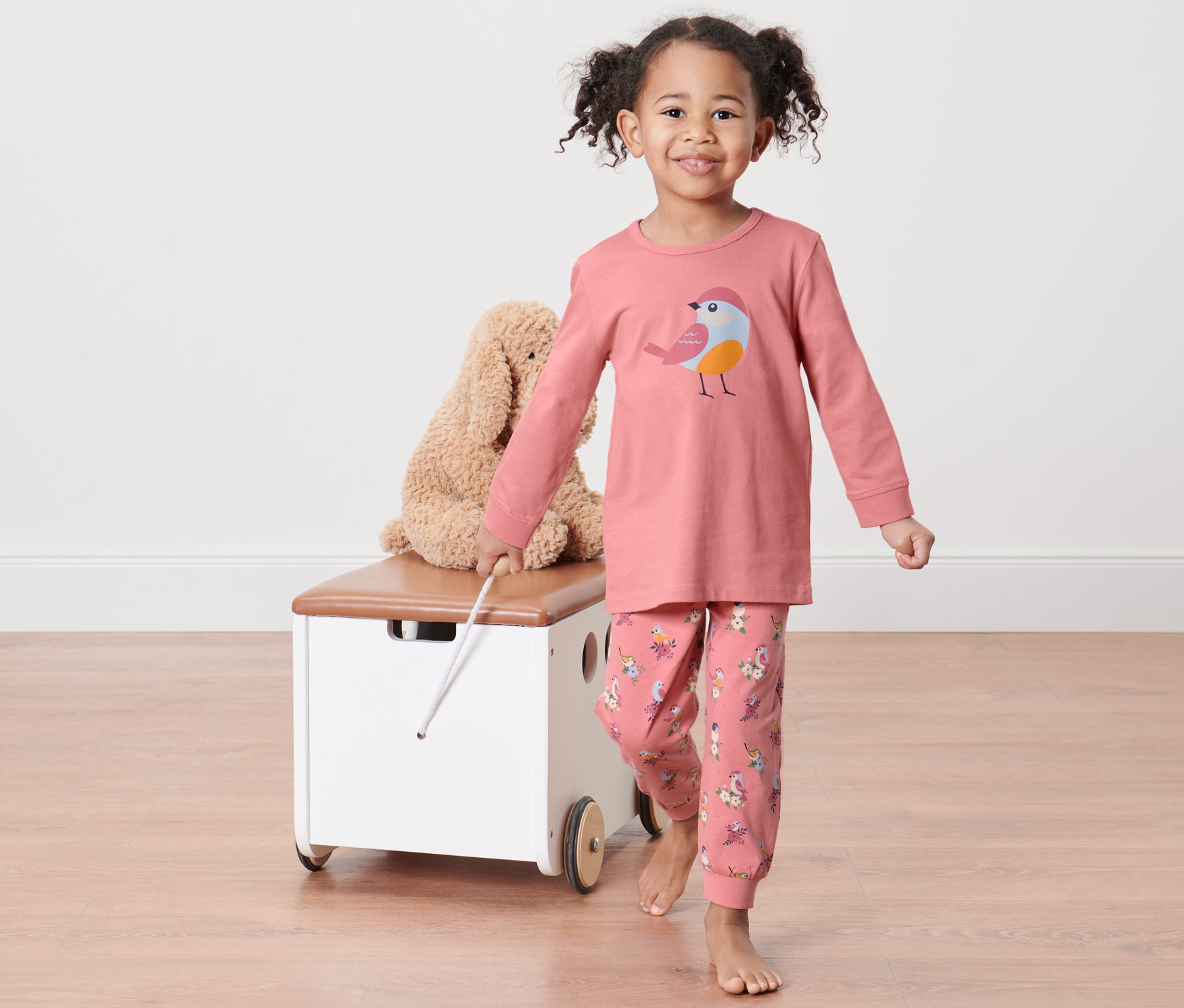 Ein Mädchen in einem 2 Kleinkind-Pyjama zieht einen Wagen mit einem Teddybär hinter sich her.