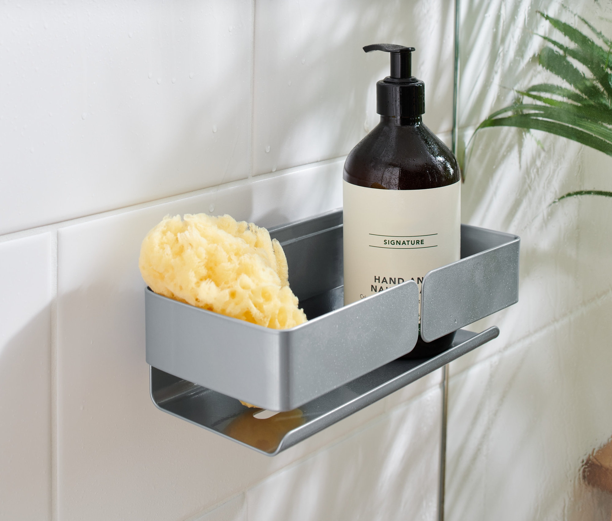 Eine graue Duschablage an weißen Fliesen hält eine braune Flasche mit Pumpspender und einen gelben Schwamm.