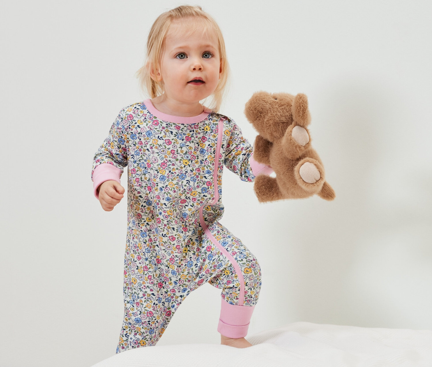 Kleinkind im geblümten Baby-Pyjama hält Teddybär, stehend auf weißer Unterlage.