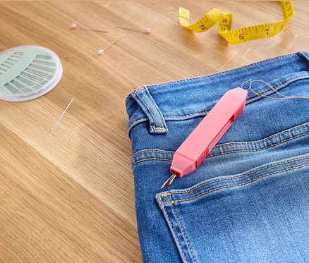 Jeans mit 2-in-1-Nähhilfe, Nadeln, Stecknadeln und gelbem Maßband auf Holzoberfläche.