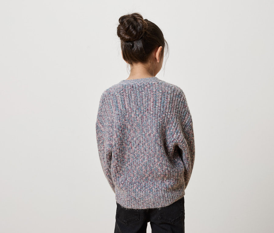 Mädchen mit Dutt steht mit dem Rücken zur Kamera und trägt einen bunten Strickpullover und schwarze Jeans.