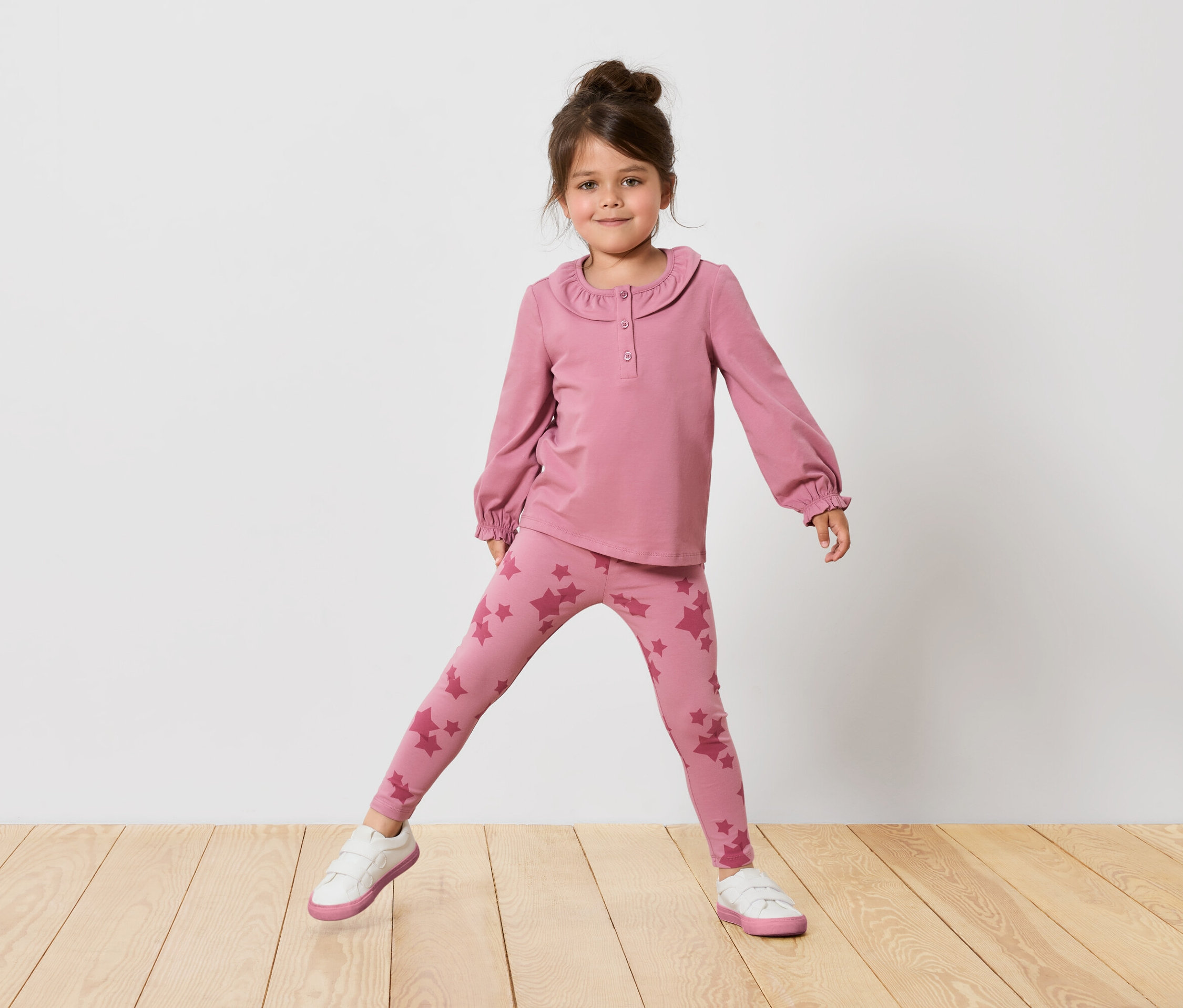 Mädchen in rosa Langarmshirt und Leggings mit Sternen steht auf einem Holzboden vor einer weißen Wand.