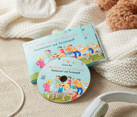 CD-Hörbuch »Wir Kinder aus dem Möwenweg – Sommerzeit und Ferienspaß« liegt mit Kopfhörern auf einer Decke neben einem Stofftier.