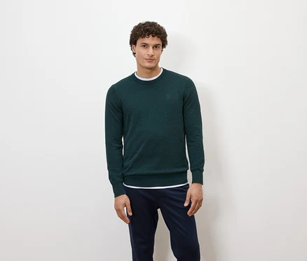Ein Mann posiert vor einem weißen Hintergrund in einem dunkelgrünen Pullover und dunkelblauen Hosen.